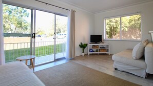 A spacious room with a large sliding glass door leading to a balcony, a comfortable white sofa, a small round table, and a television on a white stand. - The Heart of Huskisson (Huskisson)