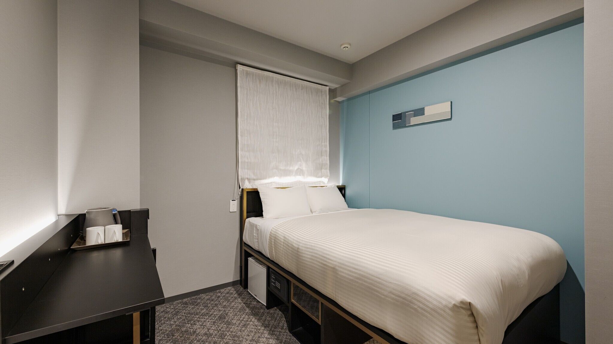 Une petite chambre d’hôtel moderne, équipée d’un lit simple, d’un bureau avec une chaise, d’une petite table avec des tasses et d’une table de chevet.