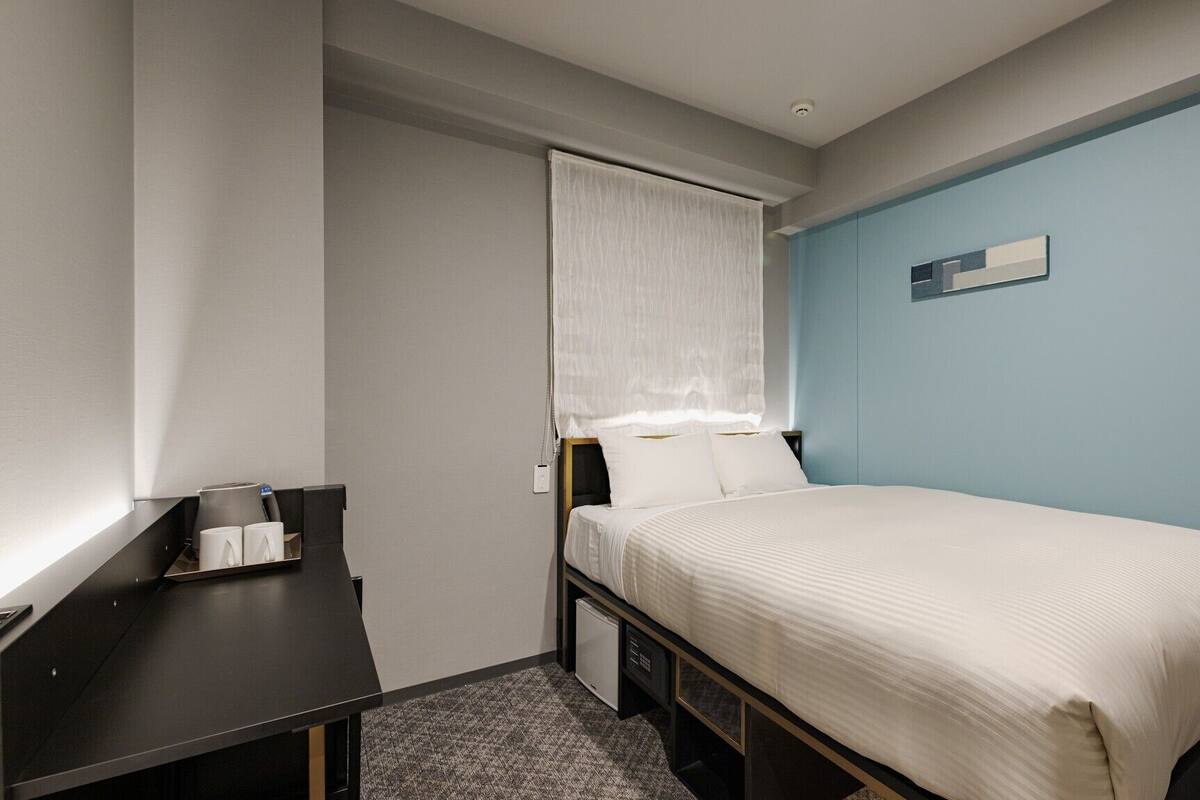 Une petite chambre d’hôtel moderne, équipée d’un lit simple, d’un bureau avec une chaise, d’une petite table avec des tasses et d’une table de chevet.