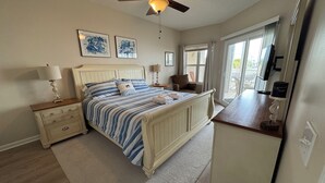A bedroom with a bed, nightstands, a ceiling fan, artwork on the wall, and a view of the outdoors. - Spacious clean condo,  large pool, plus many extras.   Perfect for families. (Oak Island)
