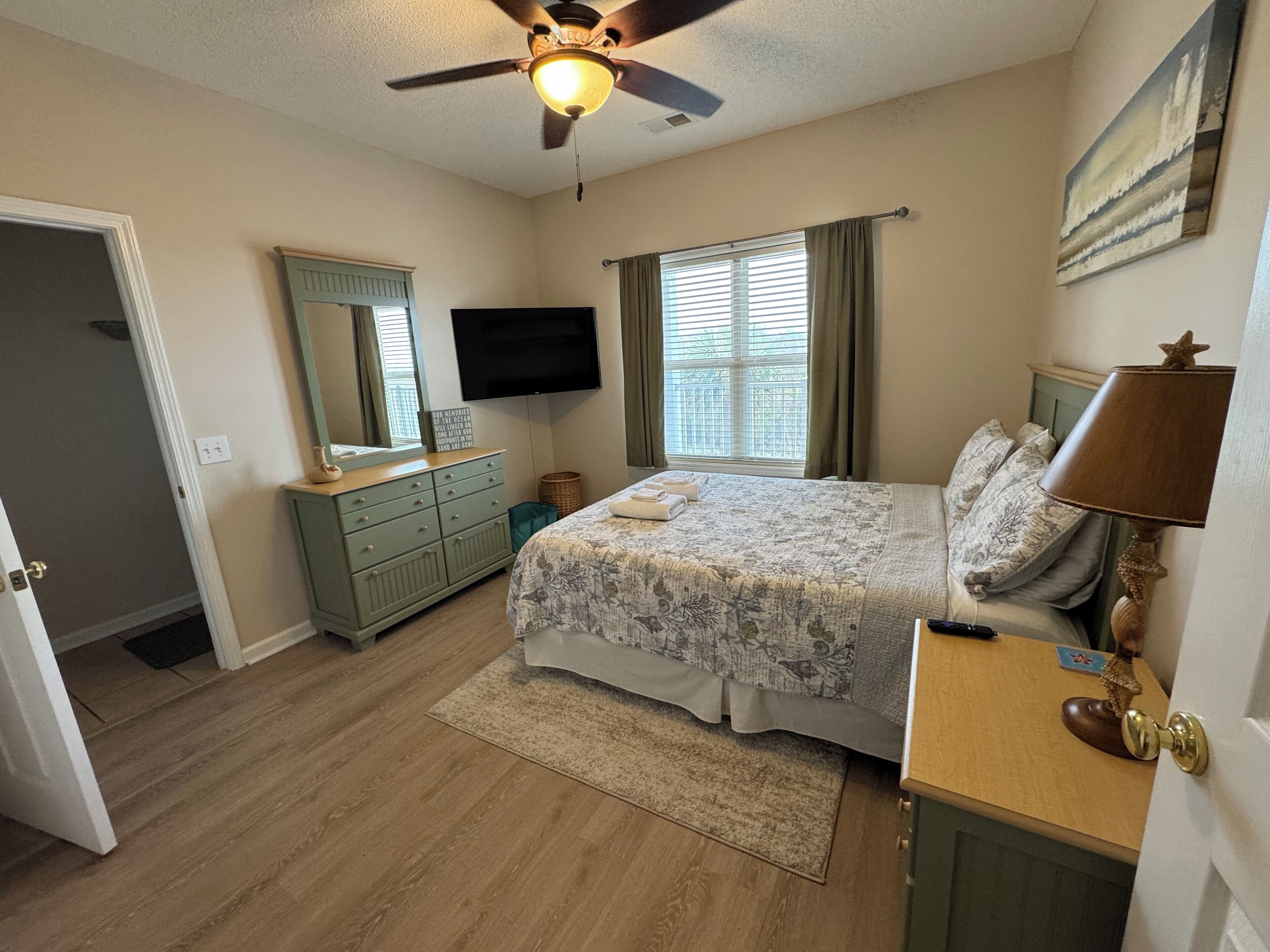 A bedroom with a bed, a television, a ceiling fan, a window with curtains, and a door.