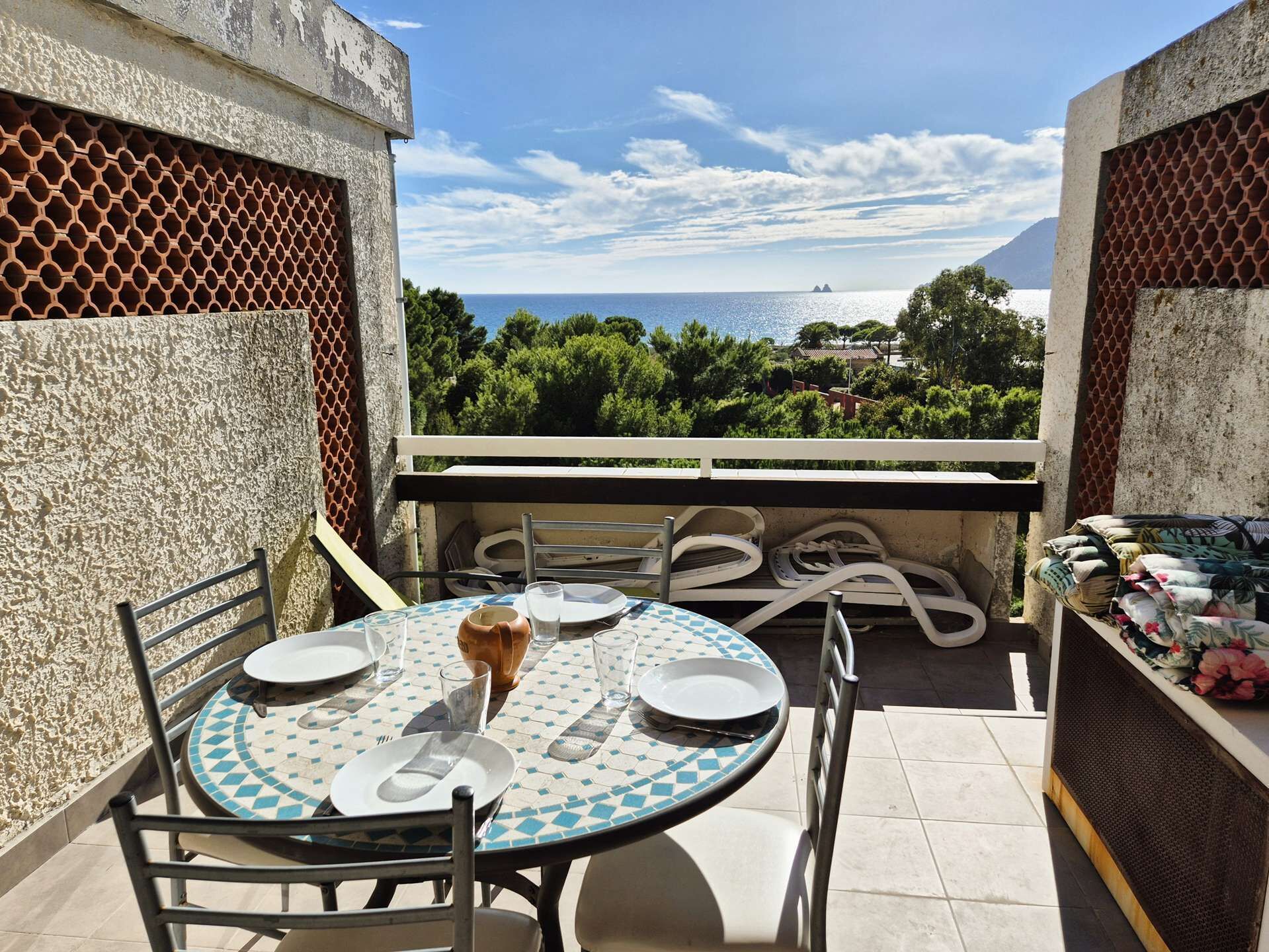 A balcony with a round table set for two, chairs, and a view of the sea and greenery.