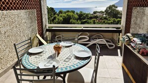 Een balkon met een ronde tafel voor twee personen, stoelen en uitzicht op zee en groen.