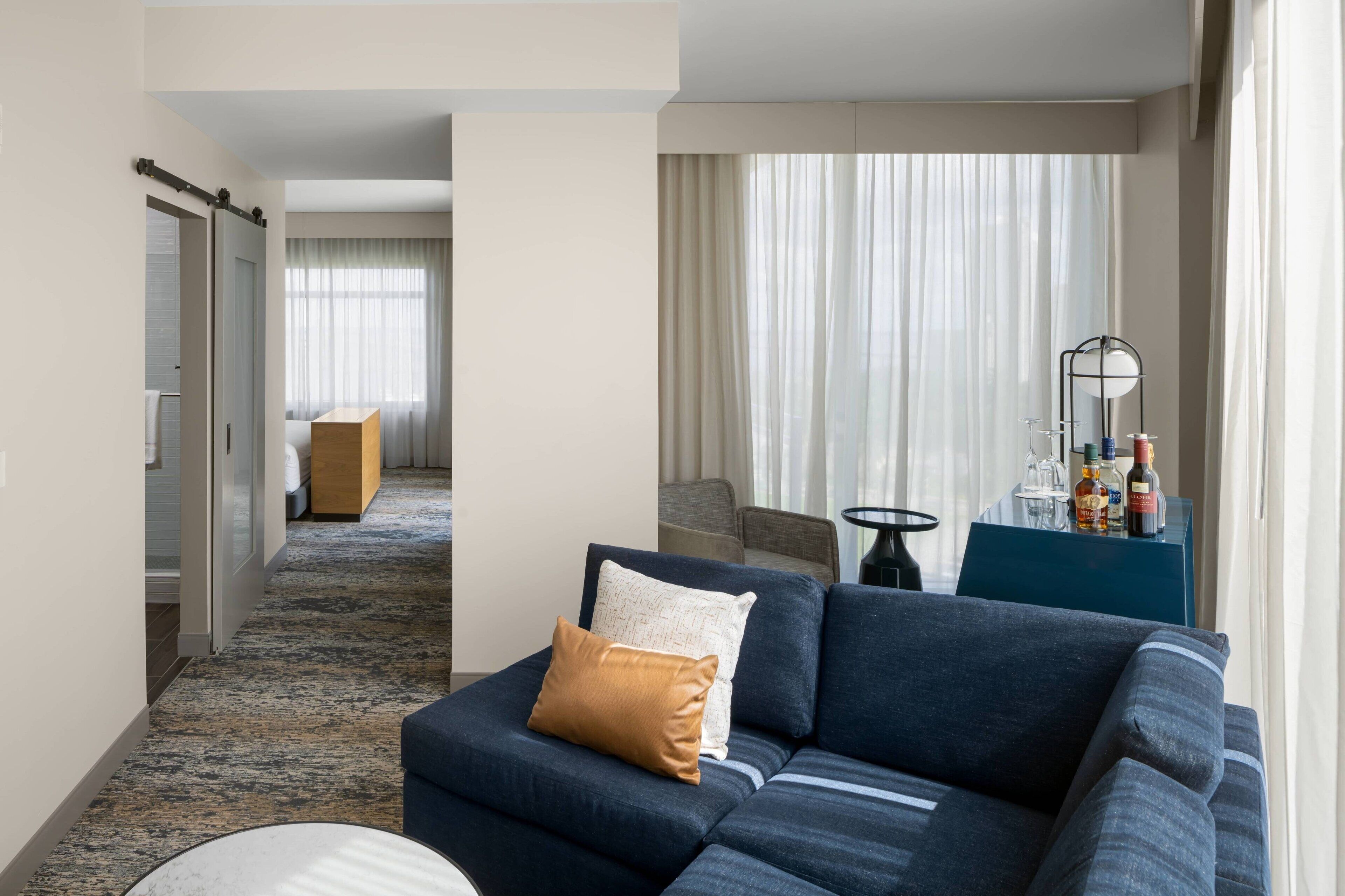A modern hotel room with a sectional sofa, a small round table, and a view of the hallway.