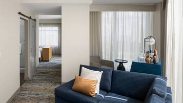 A modern hotel room with a sectional sofa, a small round table, and a view of the hallway.