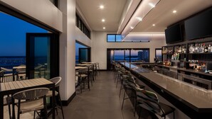 A modern bar area with a long counter, multiple seating options, and a view of the cityscape at dusk. - Luminary Hotel & Co., Autograph Collection (Fort Myers)