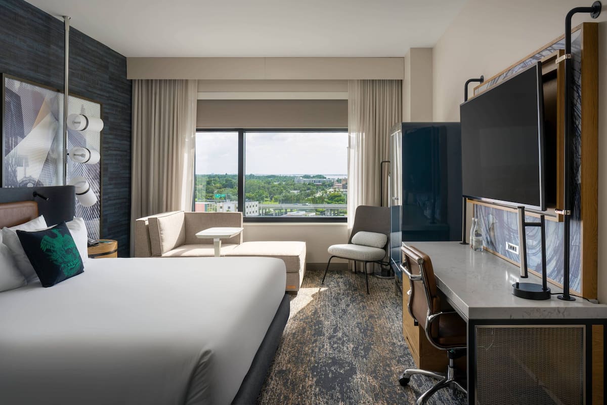 A hotel room with a large bed, a desk with a chair, a television, and a view of the city through large windows.