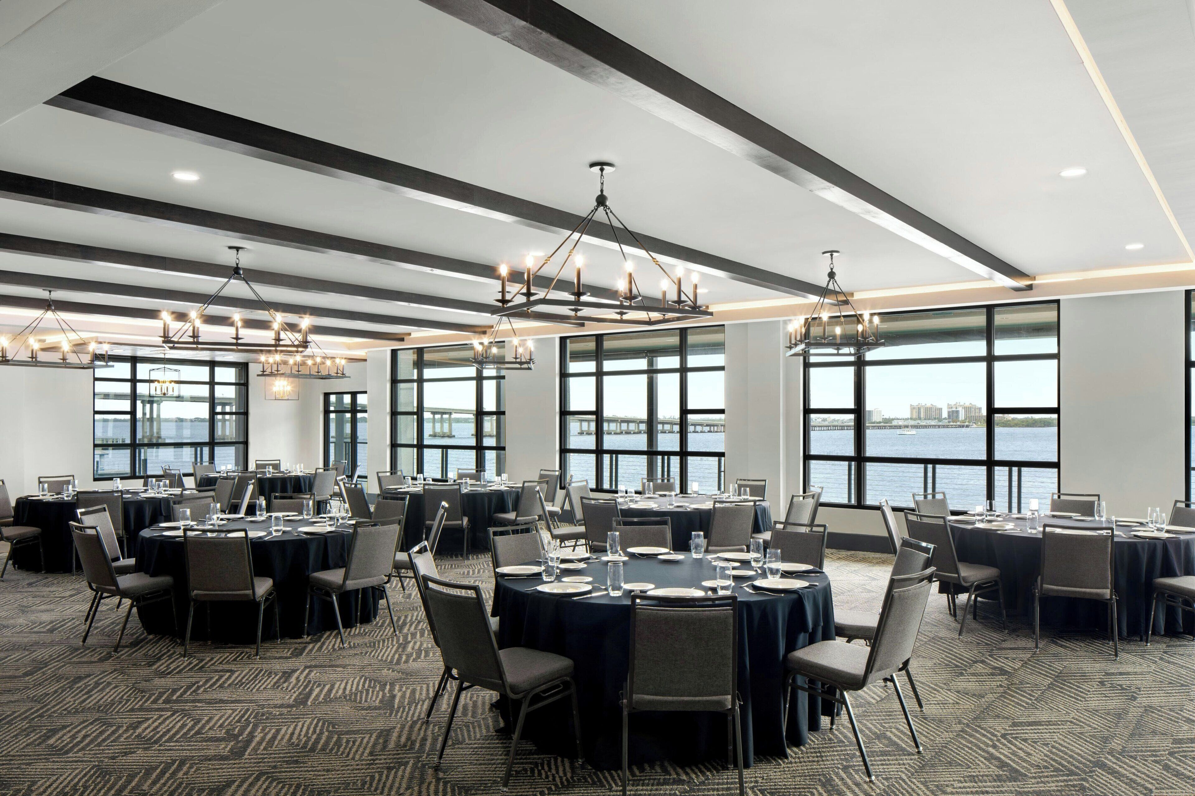 A conference room with round tables, chairs, and chandeliers. Large windows offer a view of the water and a bridge.