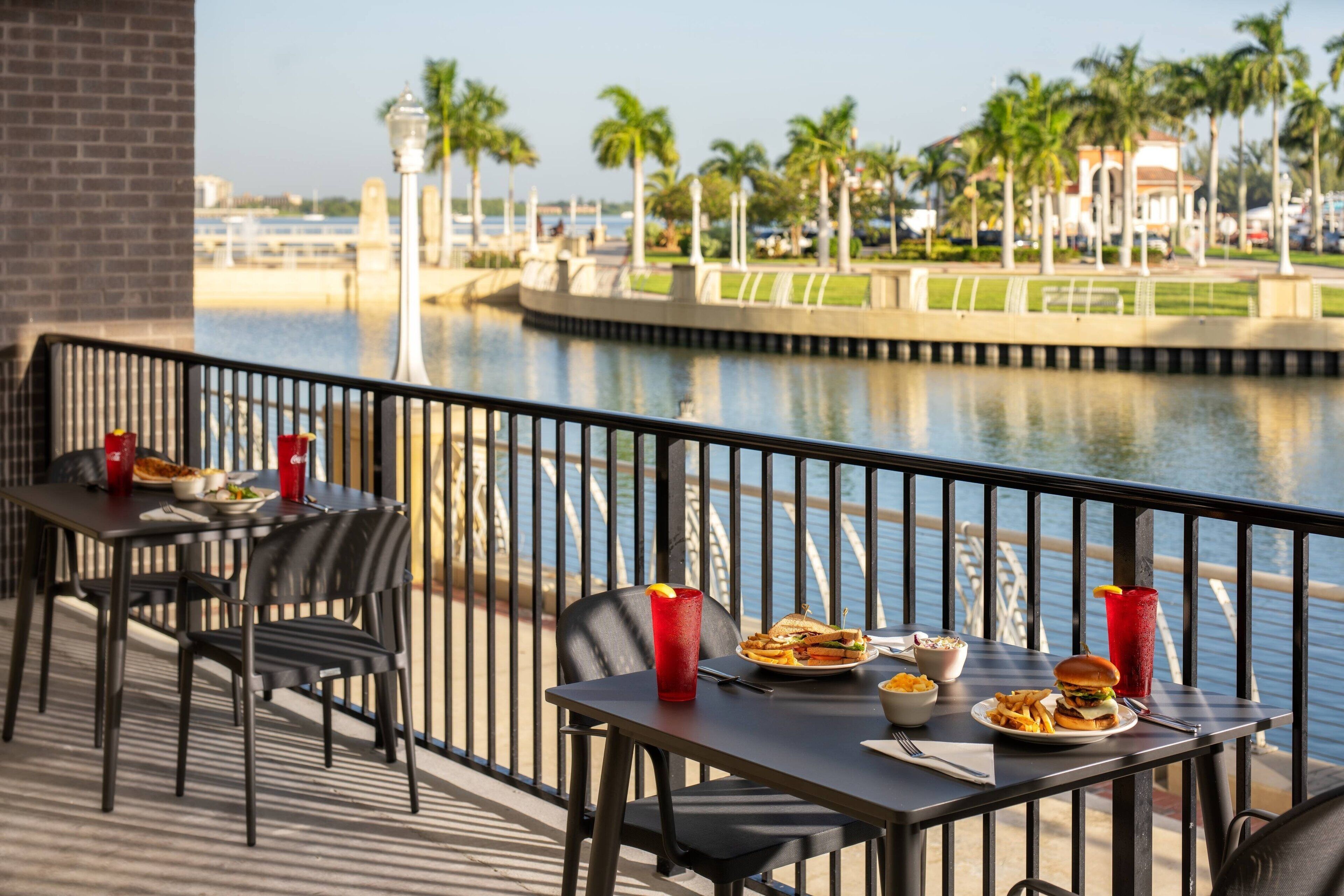 Una mesa de comedor con comida y bebidas en un balcón con vistas a un cuerpo de agua, con palmeras y un puente al fondo.