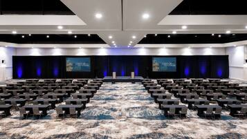 Un amplio salón de conferencias con filas de mesas y sillas, un escenario con una cortina azul y dos pantallas grandes que muestran 'CALOJA' en las paredes.