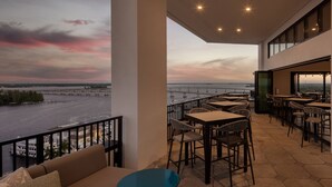 An outdoor terrace with tables and chairs, overlooking a river and a bridge. - Luminary Hotel & Co., Autograph Collection (Fort Myers)