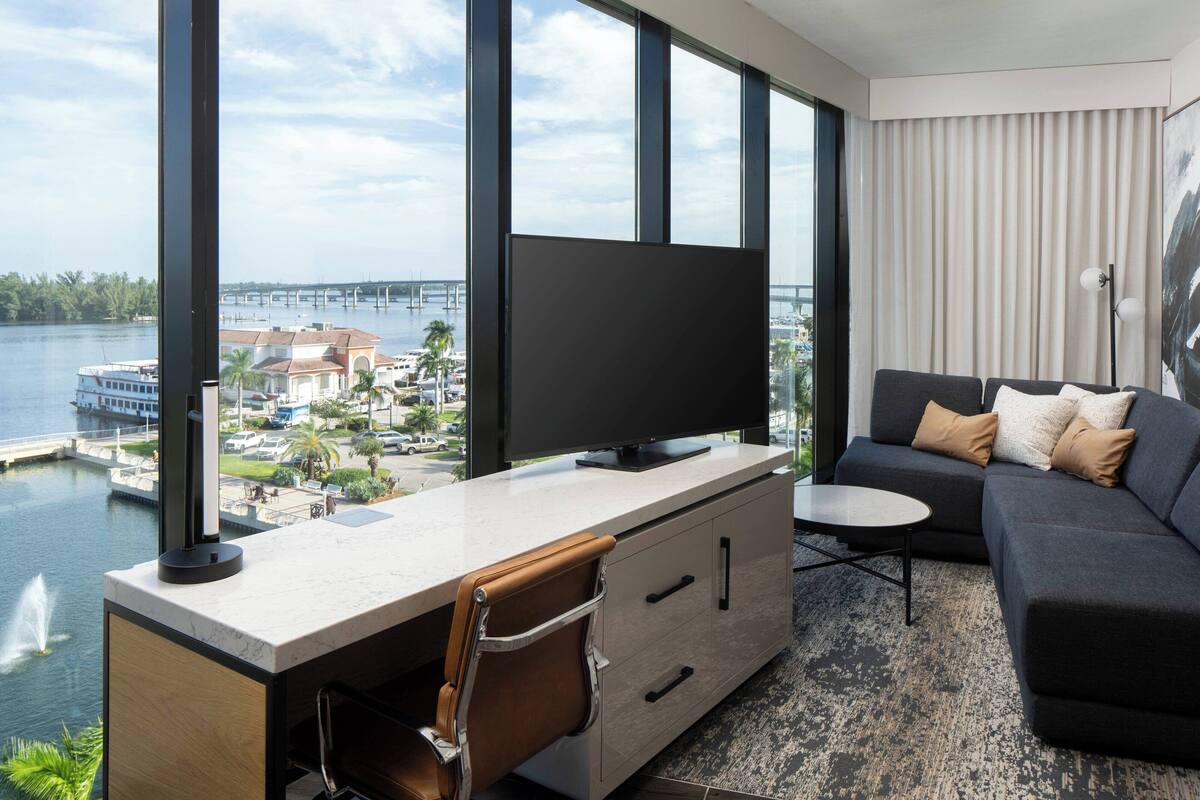 A modern hotel room with a large window offering a view of a marina and a bridge. There's a flat-screen TV on a desk, a sofa with cushions, and a small table.
