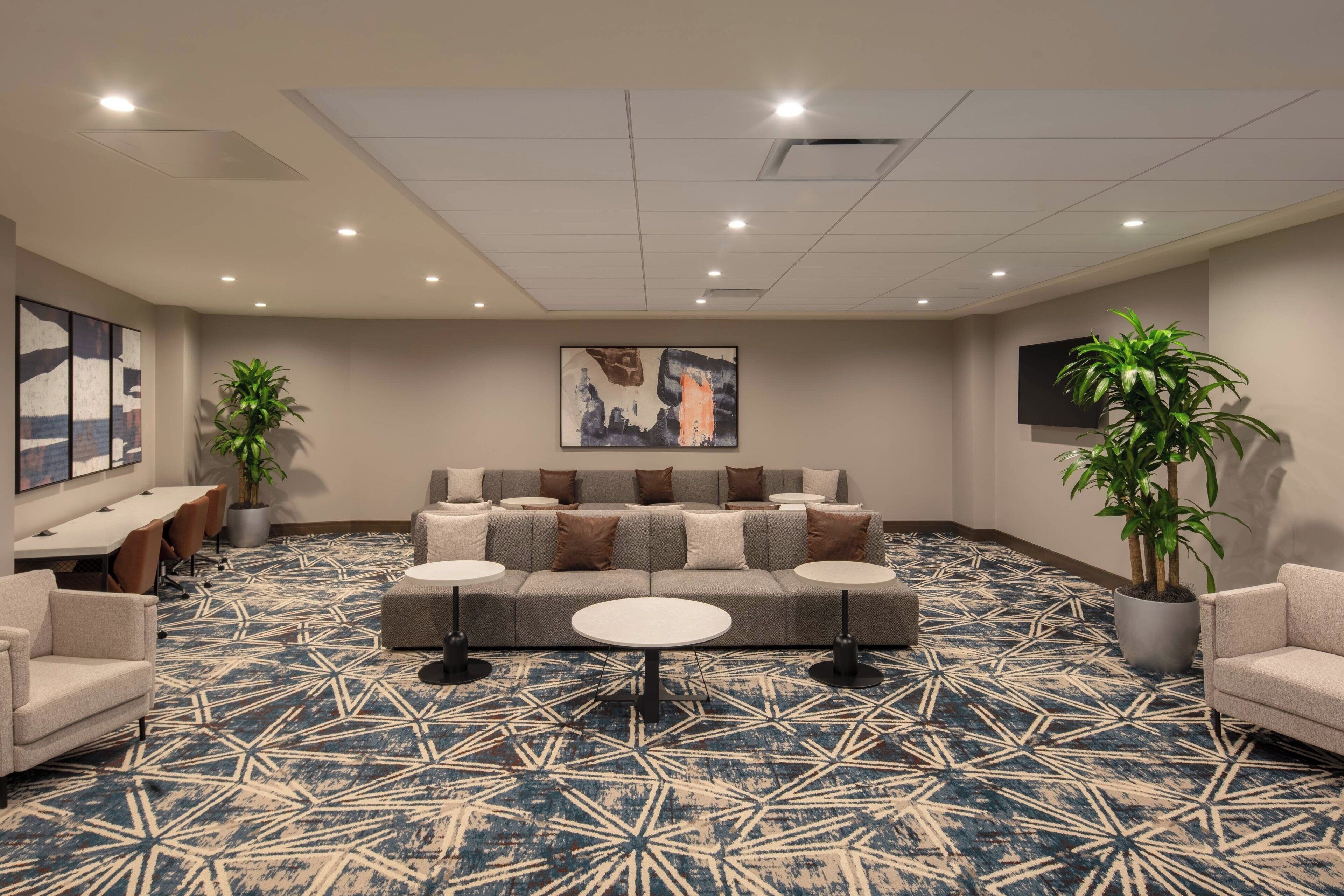 Een hotellobby met een kamerbreed tapijt, een bank met meerdere kussens, een salontafel en twee potplanten.