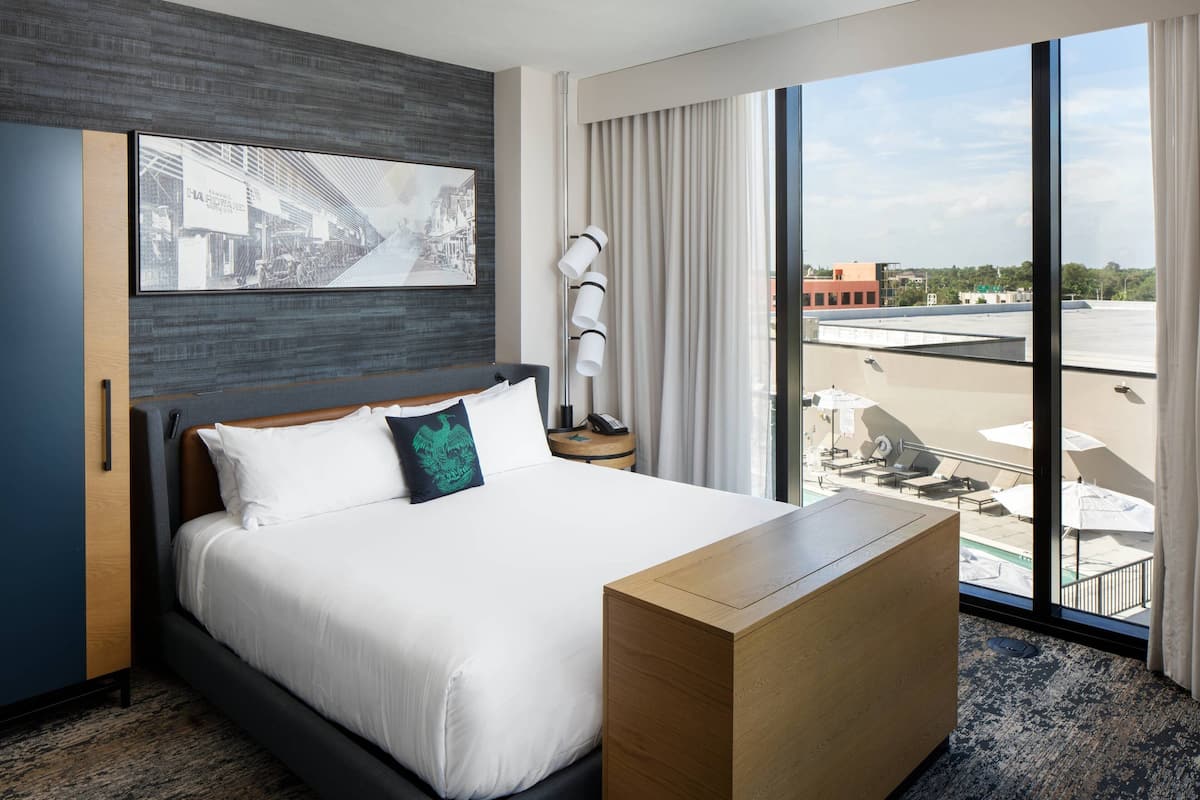 A hotel room with a large bed, a wooden desk, a nightstand, a framed picture on the wall, and a view of the city through large windows.