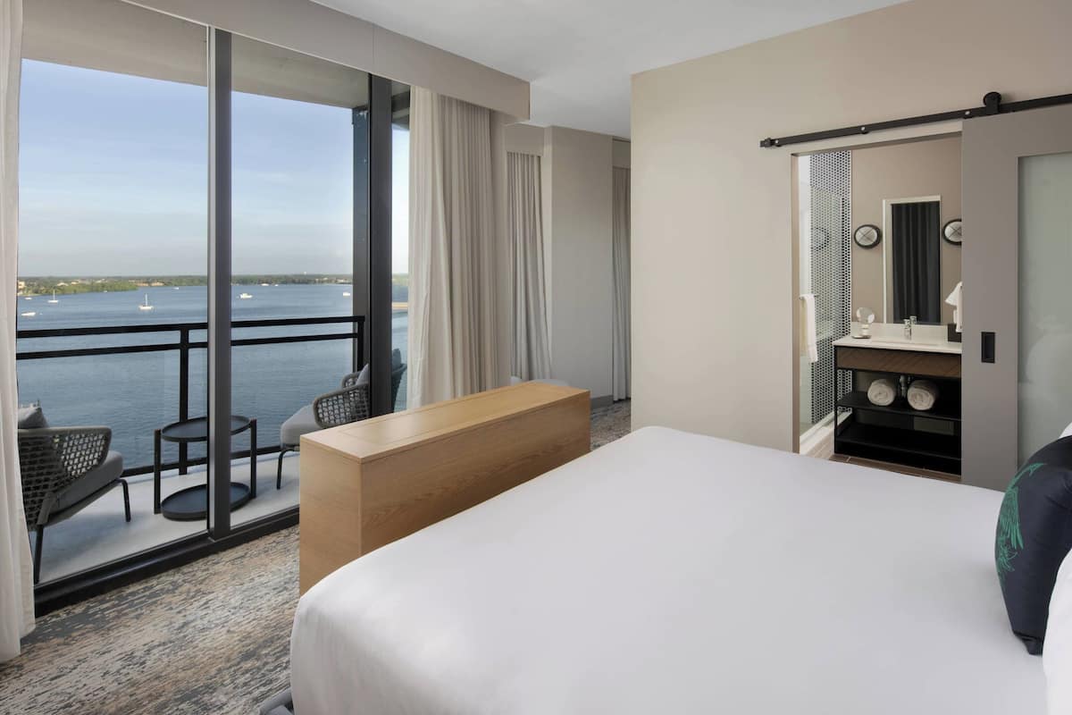 Habitación de hotel con una cama grande, un escritorio de madera, una zona de estar con sillas, un baño con espejo y lavamanos, y vistas al agua.