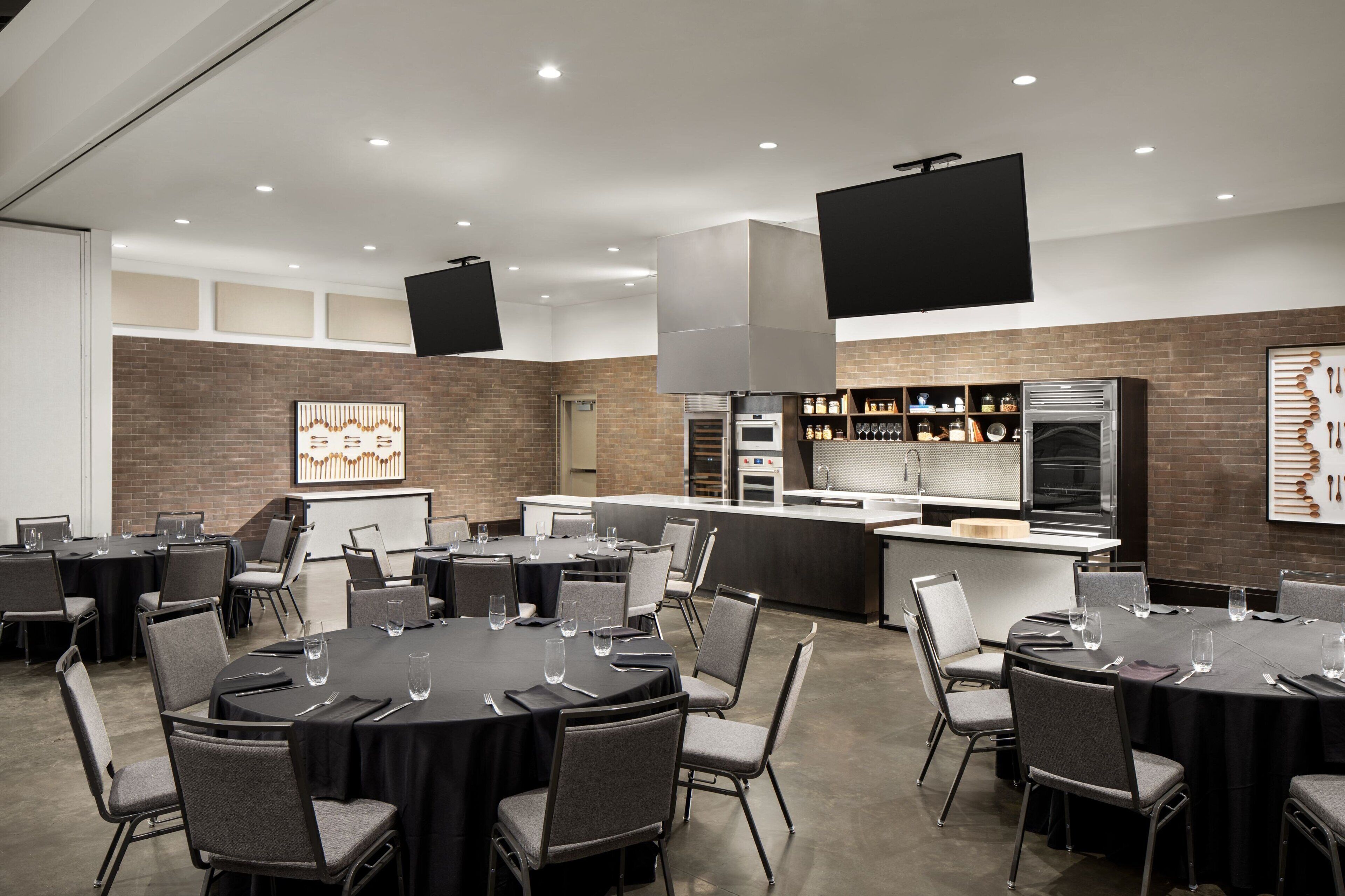 A modern conference room with round tables, chairs, a kitchen area, and a large flat-screen TV.