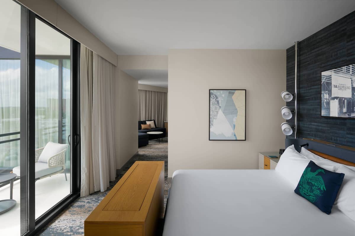 A hotel room with a large bed, a wooden bench, a balcony with a chair, and a view of the city.
