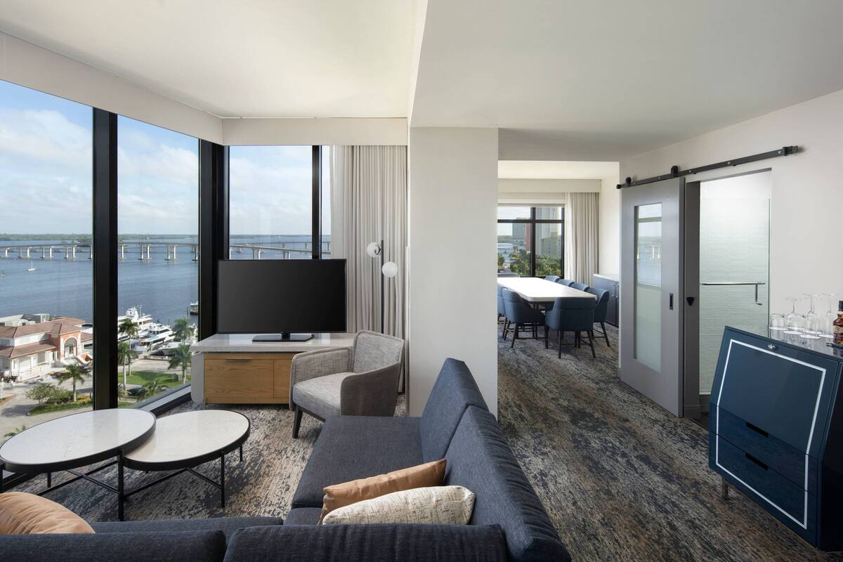 A modern hotel room with a large window offering a view of the sea and a bridge. There is a sofa, a small table, and a TV on a wooden cabinet.