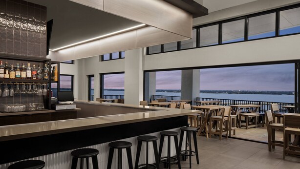 A modern bar area with a long counter, stools, and a view of the water. - Luminary Hotel & Co., Autograph Collection (Fort Myers)