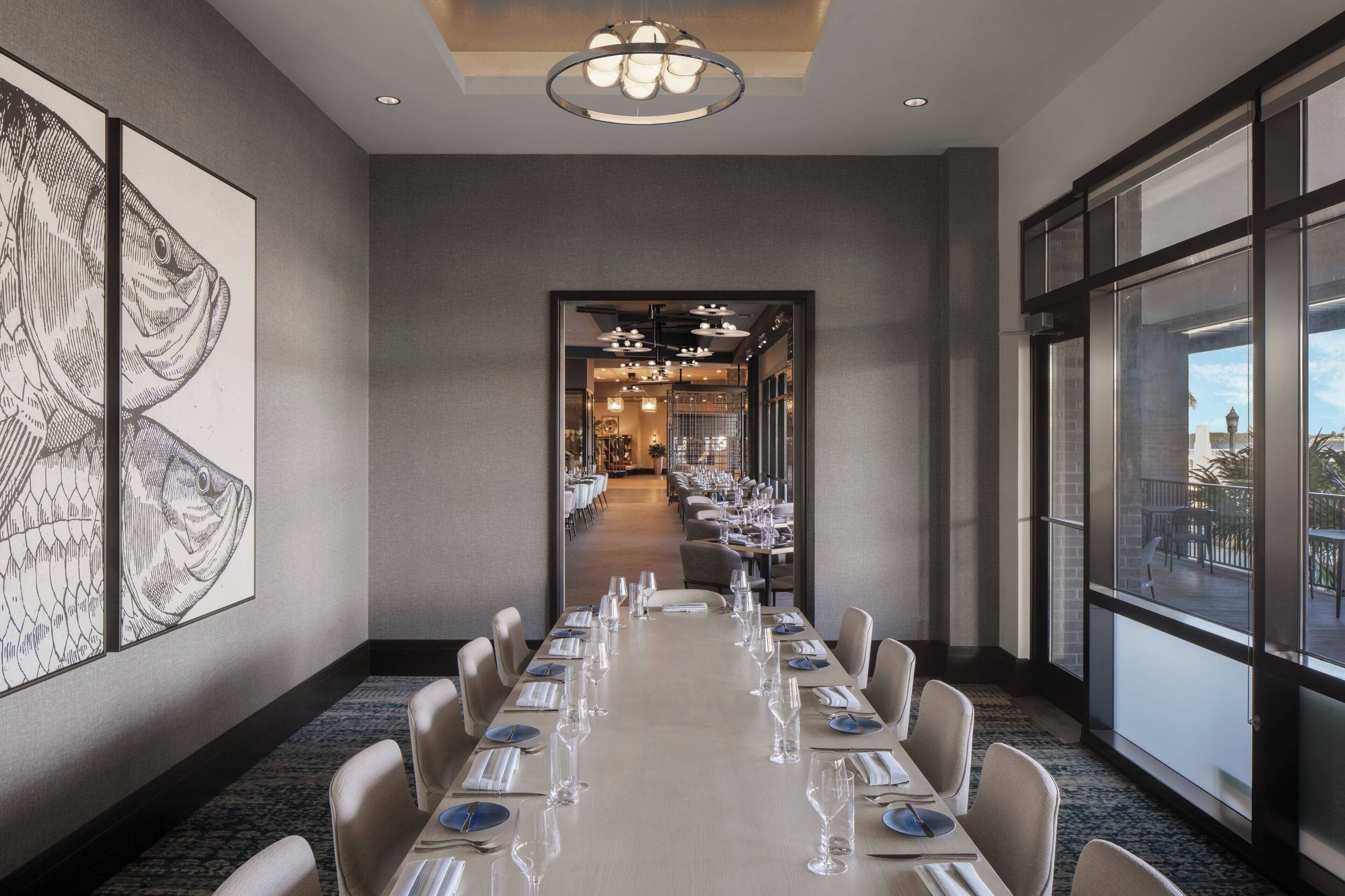 Une longue table à manger, dressée pour un repas, se trouve dans une chambre d’hôtel moderne dotée de grandes fenêtres et ornée d’œuvres d’art sur les murs.