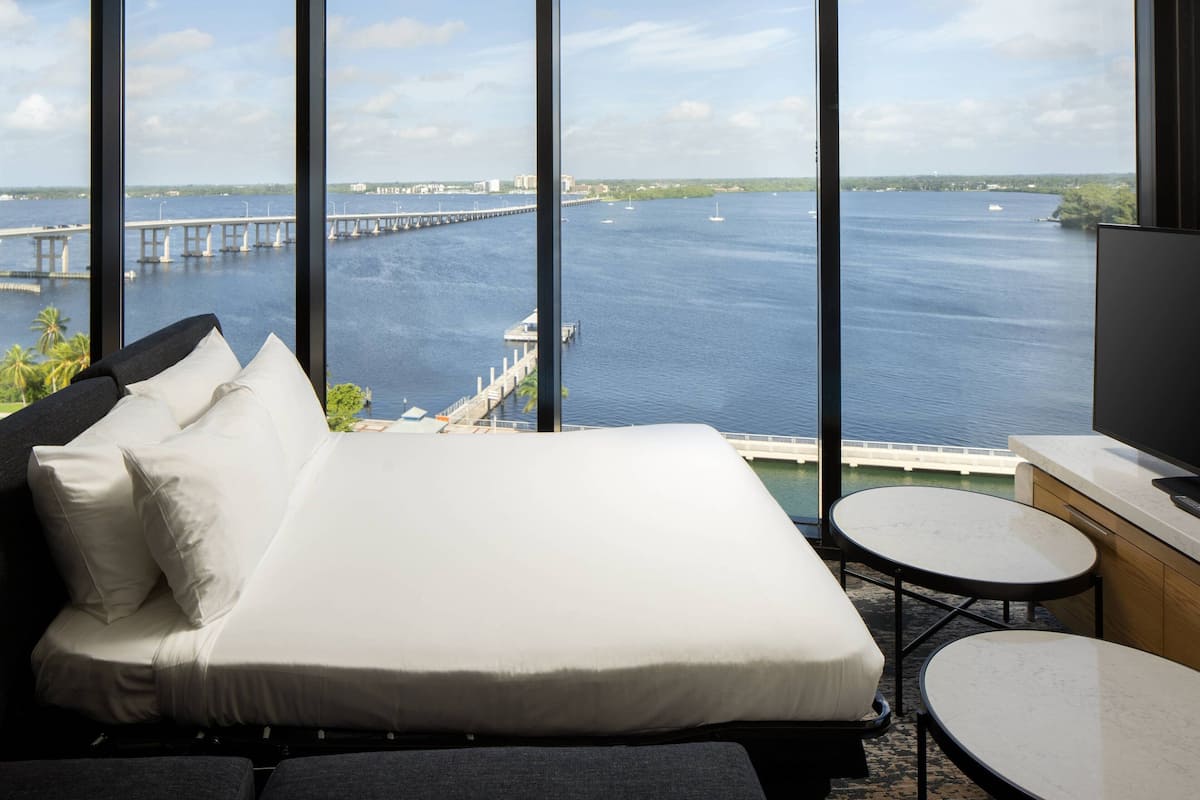 Habitación de hotel con una cama grande, una mesita redonda y un televisor. Ofrece vistas de una masa de agua con un puente y barcos.