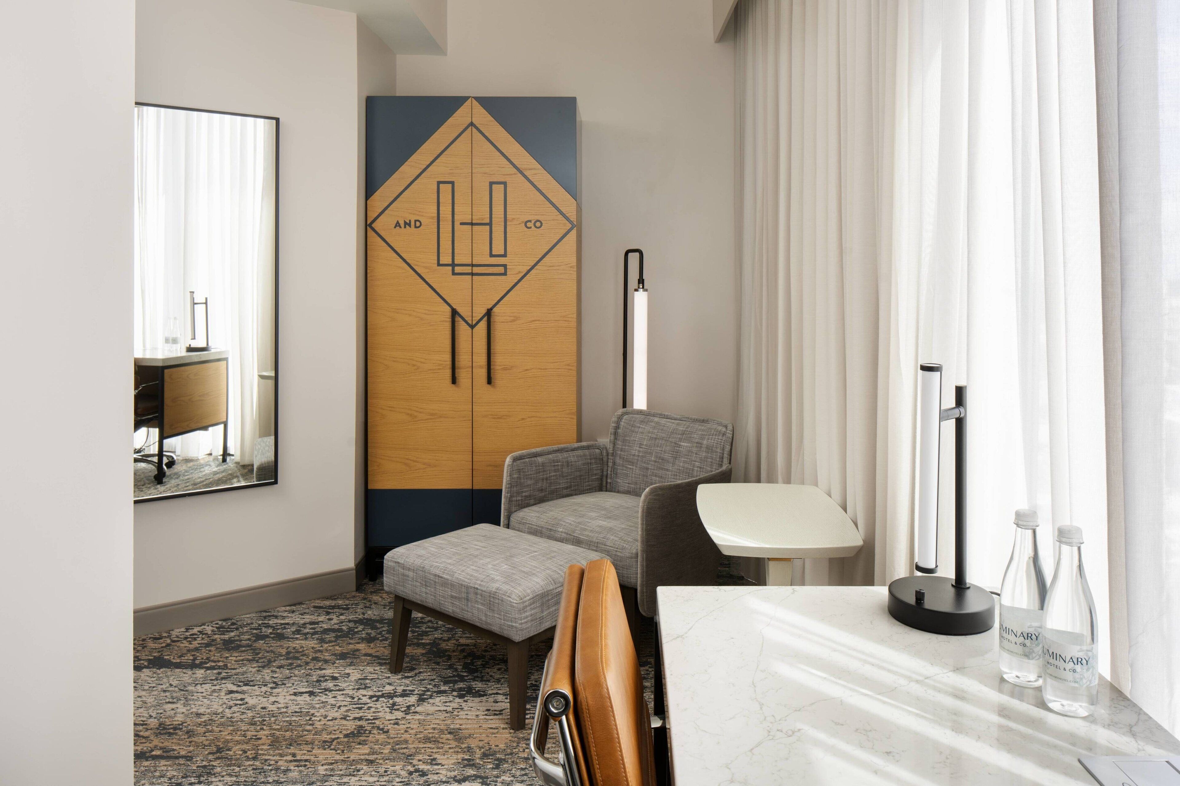 A hotel room with a large mirror, a wooden accent wall with a geometric design, a gray upholstered chair, a small table with a lamp, and two bottles of water.