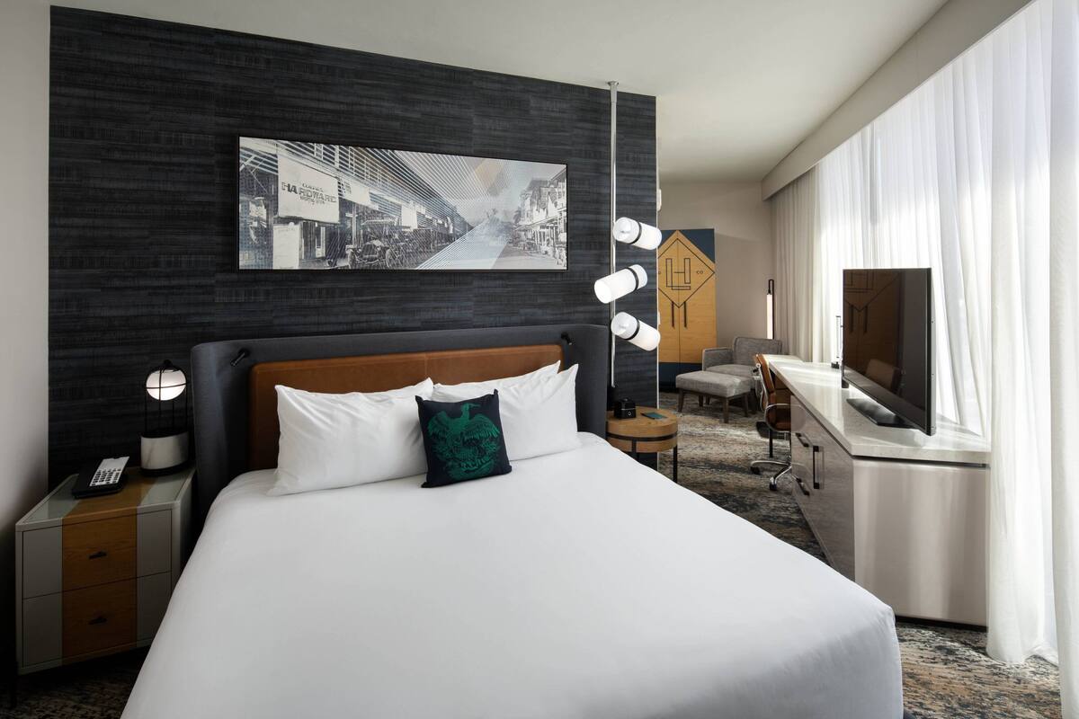 A hotel room with a large bed, a desk, a chair, a TV, and a painting on the wall.