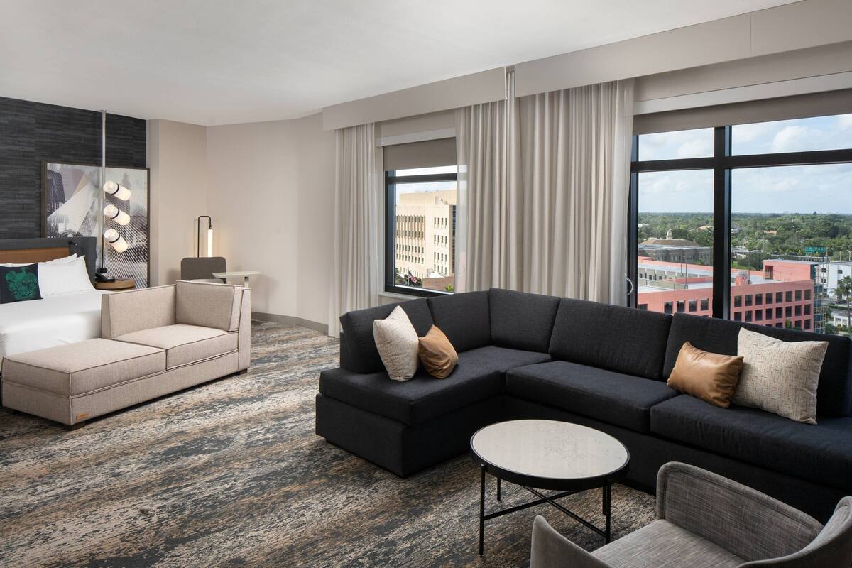A modern hotel room with a sectional sofa, a separate sofa, a round coffee table, and a large window offering a city view.