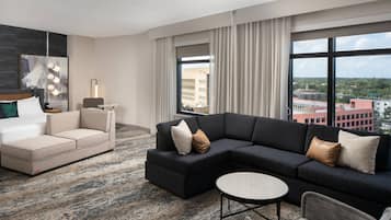 Et moderne hotelværelse med en hjørnesofa, en enkelt sofa, et rundt sofabord og et stort vindue med udsigt over byen.
