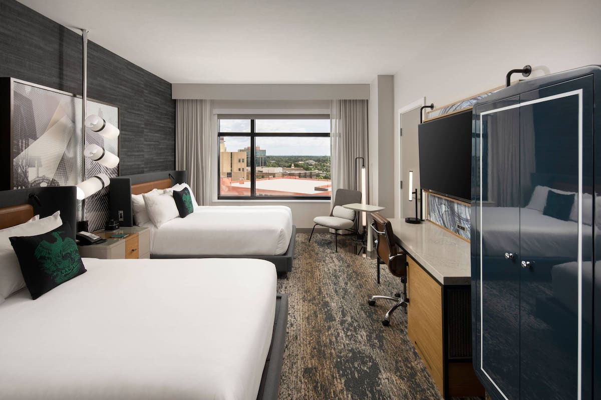 A hotel room with two beds, a desk, a chair, a TV, and a large window offering a city view.