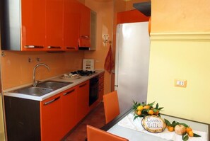 Uma cozinha compacta com armários cor de laranja, uma geladeira branca e uma pequena mesa de jantar com um centro de frutas.