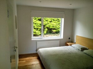 A bedroom with a bed, a nightstand, a window with a view of greenery, and a radiator. - Exclusive Garden Apartment (Galway)