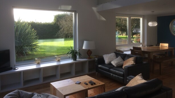 A spacious living room with a large window, a black leather sofa, a wooden coffee table, and a dining area with a table and chairs. - Exclusive Garden Apartment (Galway)