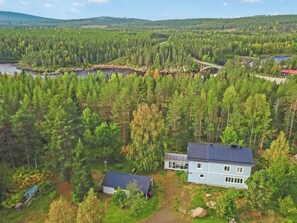 Et stort hus med mørkt tag, omgivet af tæt skov og et mindre vandareal.