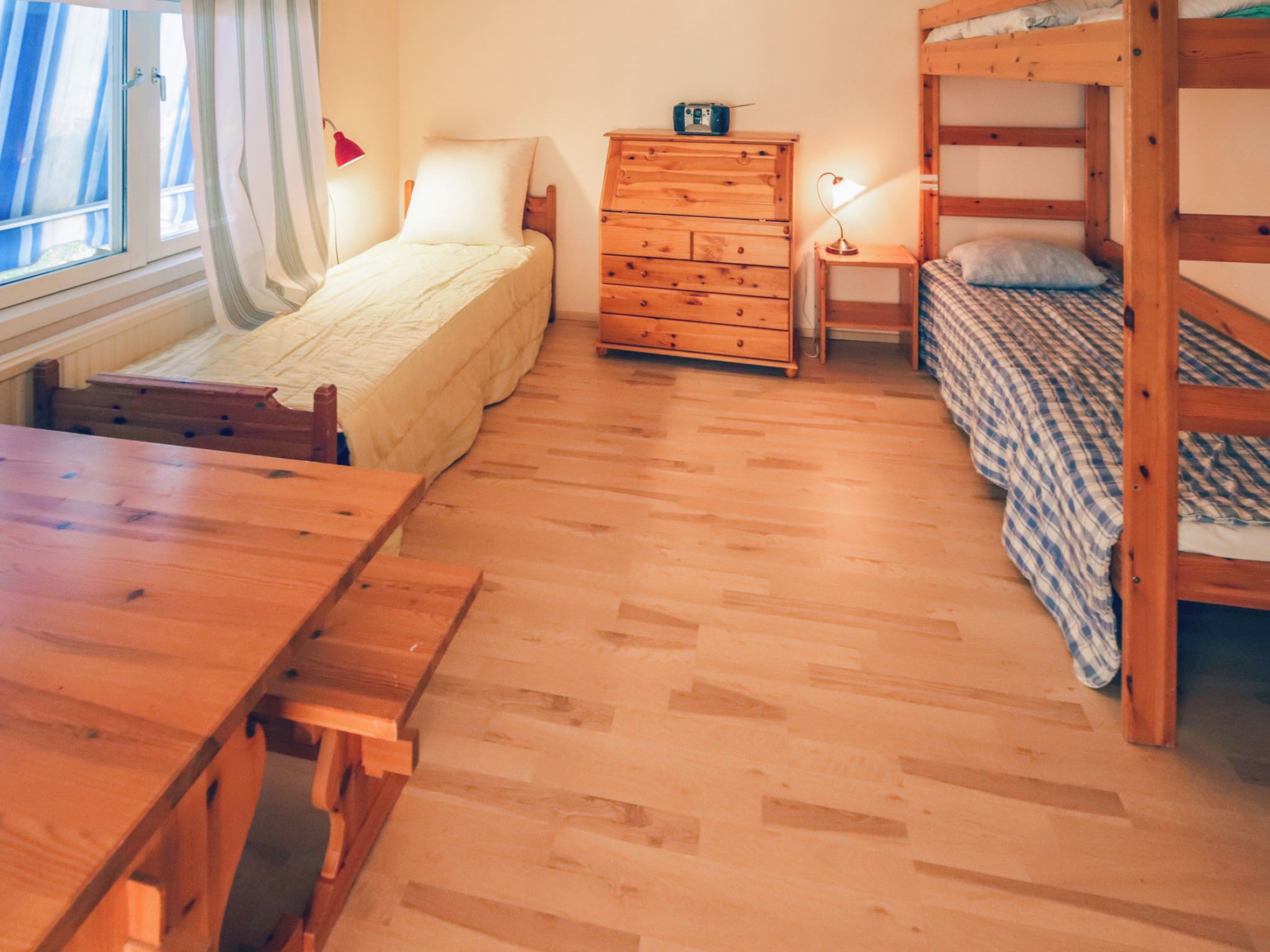 Une pièce de taille modeste comprenant un lit superposé, un lit simple, une commode en bois, un bureau et une fenêtre avec des rideaux.