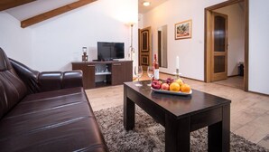 Un salon avec un canapé en cuir marron, une table basse en bois foncé sur laquelle sont disposés des fruits et des verres à vin, un téléviseur posé sur un meuble en bois, et une poutre en bois au plafond.