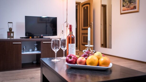 Un coin cuisine avec une table à manger dressée, agrémentée de fruits, de verres à vin et d’une bouteille. On aperçoit un téléviseur au fond.