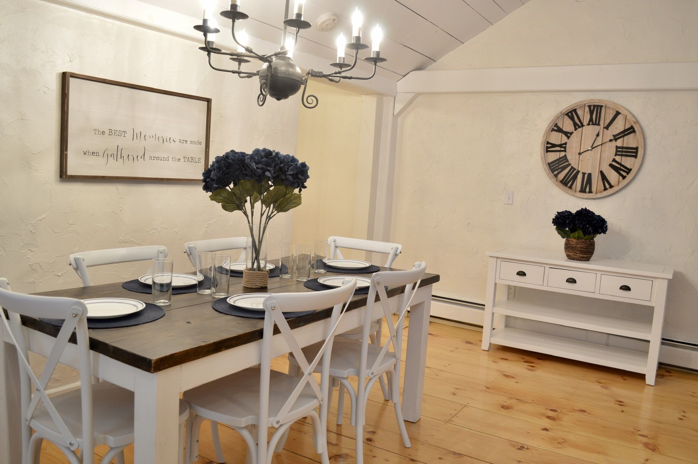 Un comedor con una mesa de madera dispuesta para cuatro, sillas blancas, una araña, un reloj de pared y una cita enmarcada.