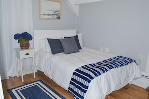 Um quarto com uma cama branca, um cobertor listrado azul e branco, uma mesa de cabeceira branca com um vaso de flores e um quadro na parede.