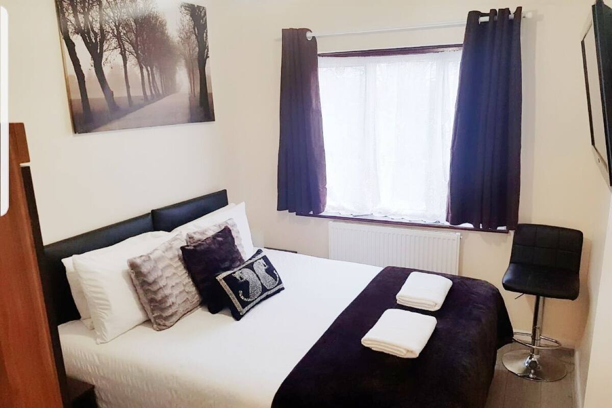 Cama bem arrumada com edredom branco e duas almofadas, manta azul escura e duas toalhas dobradas. Há uma janela com cortinas escuras, um quadro com uma trilha em uma floresta na parede e um abajur preto ao lado da cama.