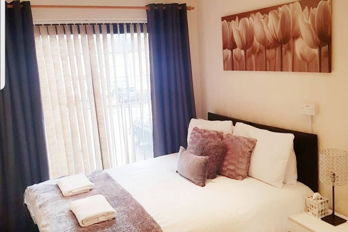 Quarto de hotel com uma cama grande, uma mesa de cabeceira com abajur e uma janela com cortinas.