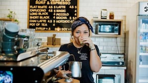 Un barista derrière le comptoir d’un café, avec un panneau affichant différentes sortes de boissons au café en arrière-plan.