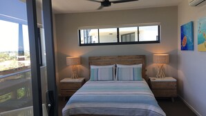 Une chambre à coucher avec un grand lit, deux tables de chevet, un ventilateur de plafond et une fenêtre donnant sur des immeubles.
