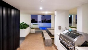 Un espace extérieur moderne, doté d’un barbecue, de sièges et offrant une vue sur une piscine au crépuscule.