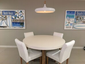 Une table de salle à manger ronde, entourée de quatre chaises blanches, un luminaire suspendu et deux œuvres d’art murales sur le thème de la plage.