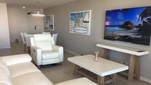 Un salon avec un canapé en cuir blanc, un fauteuil blanc et une table basse. Deux tableaux sont accrochés au mur, et un téléviseur à écran plat repose sur un meuble en bois.