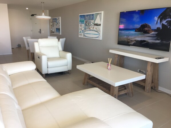 Un salón con un sofá de cuero blanco, un sillón blanco y una mesa de centro. Hay dos cuadros colgados en la pared y un televisor de pantalla plana sobre un soporte de madera.