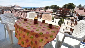 Un balcón con una mesa cubierta con mantel de estampado de manzanas, sillas blancas de plástico y una botella de vino.