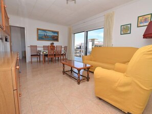 A living room with a yellow sofa, wooden dining table, and chairs. There is a painting on the wall and a balcony with a view.
