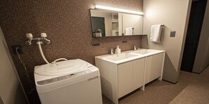 Een hotelbadkamer met een wastafel met twee kommen, een spiegel, een handdoekrek en een wasmachine.